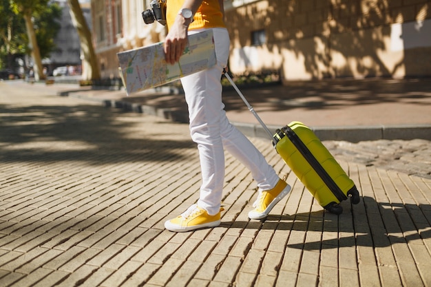 Cropped image of traveler tourist woman in yellow casual clothes with suitcase city map walking in city outdoor. Girl traveling abroad to travel on weekends getaway. Tourism journey lifestyle concept.