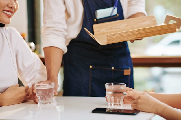 Обрезанное изображение официанта, ставящего стаканы с пресной водой перед молодыми женщинами за столиком в кафе