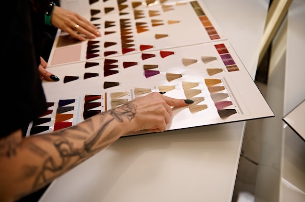 Cropped image of a well-groomed tattooed hairdresser arm pointing to a swatch of straight dyed hair in a hair dye color palette catalog and advising a client on hair coloring in beauty salon. Close-up