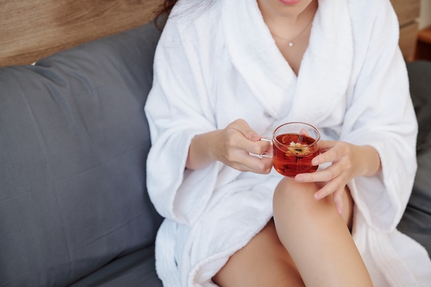 Cropped image of woman in bathrobe sitting on sofa with cup of tasty herbal tea