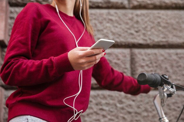 Cropped image of woman dressed in sweater walking with her bicycle in the city while standing near wall and listen to music by her phone