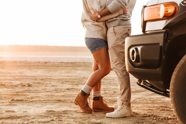 Immagine ritagliata di una giovane coppia in piedi vicino a un'auto sulla spiaggia assolata, abbracciata