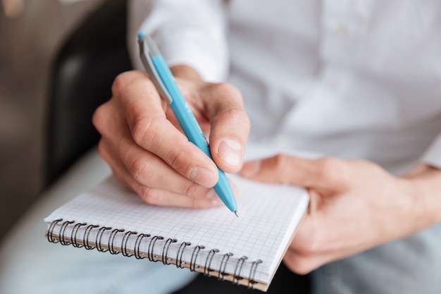 Cropped image of young man dressed in white shirt writing notes in notebook. Coworking.