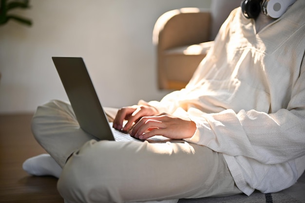 Обрезанное изображение молодого человека, сидящего на полу в гостиной и опирающегося на диван с помощью ноутбука