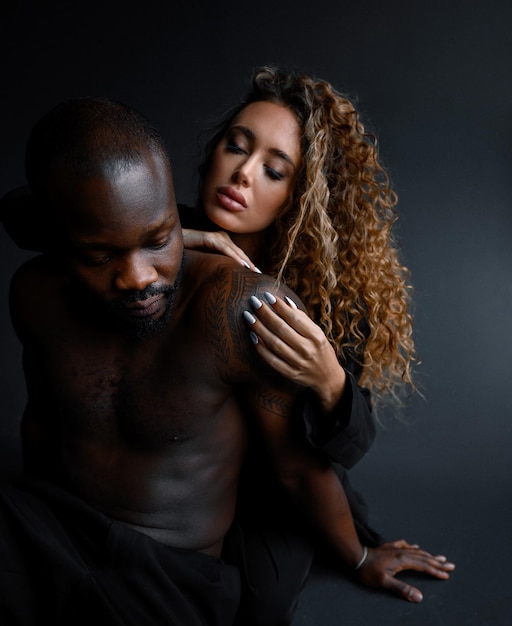 Снимок влюбленной пары: темнокожий мускулистый мужчина в брюках сидит и смотрит вниз, в то время как красивая женщина с афро-прической и белым маникюром обнимается за спиной и позирует в помещении