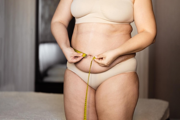 Cropped photo of fat plump woman standing in beige bra underpants showing excessive belly measuring waist using tape