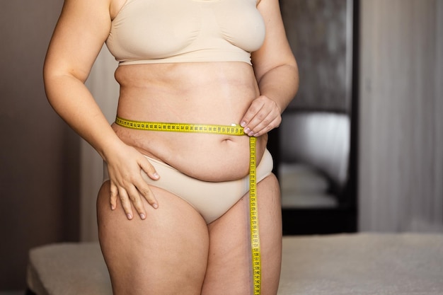 Cropped photo of fat plump woman standing in beige bra underpants showing naked belly measuring waist using tape
