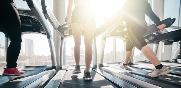 Cropped photo of female legs in sportswear and sneakers running on a treadmill in the gym against the window