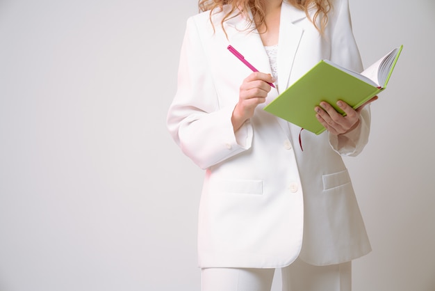 Cropped photo of a girl in white suit that holds pink pen and green notebook in her hands isolated