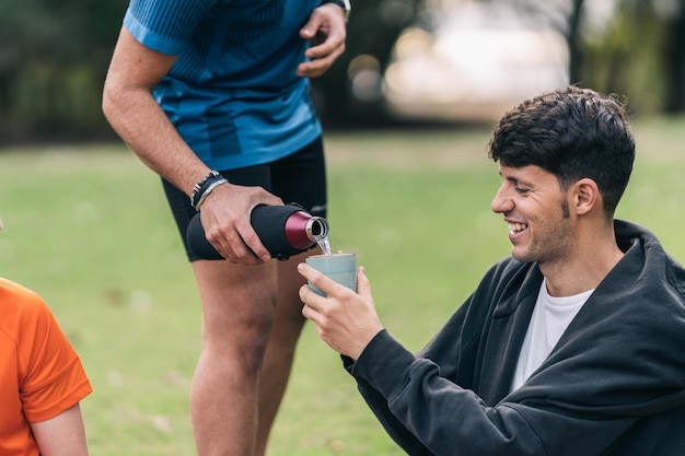 Photo cropped photo of a man pouring tea from a thermos to an sportive smiling man sitting in a park