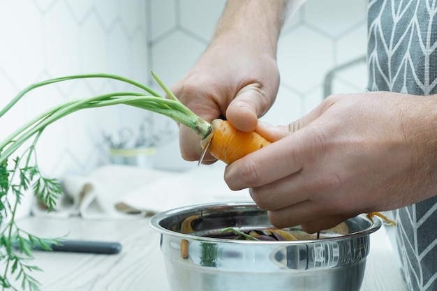 Cropped photo of man wearing grey apron cutting off leaves at top of carrot with knife over metal bowl full of peelings