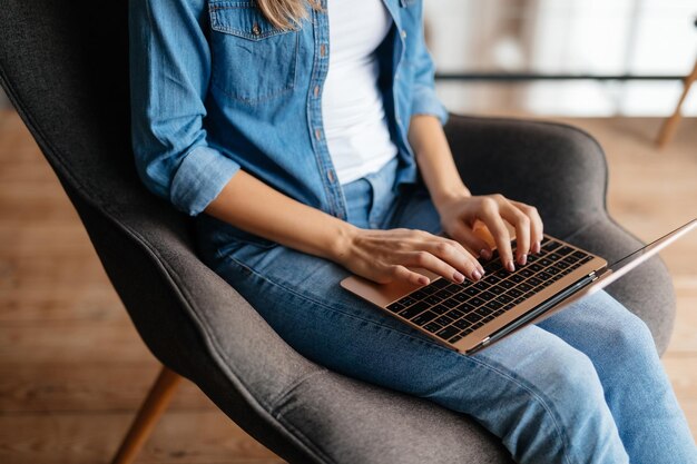 Cropped photo of slender young woman typing on keyboard of her laptop while sitting in armchair