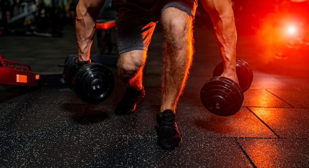 Обрезанное фото сильного атлетичного мужчины, берущего гантели с пола, тренирующегося в современном тренажерном зале Теплый оранжевый свет