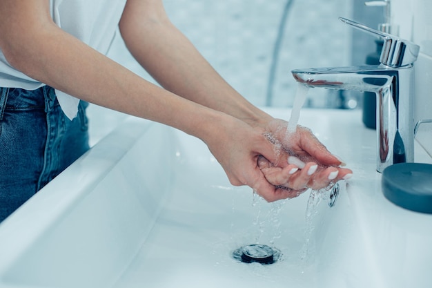 Обрезанное фото до неузнаваемости женщины, кладущей руки под чистую проточную воду в раковине ванной комнаты