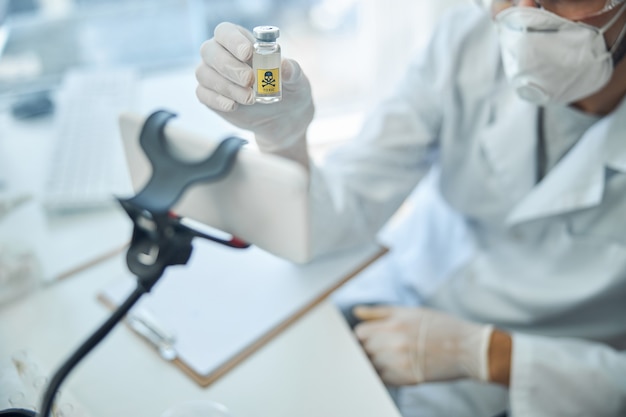 Cropped photo of a virologist in a protective mask showing a toxic substance to his online audience