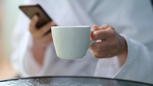 Foto ritagliata di una donna in accappatoio di spugna con in mano un telefono cellulare e una tazza