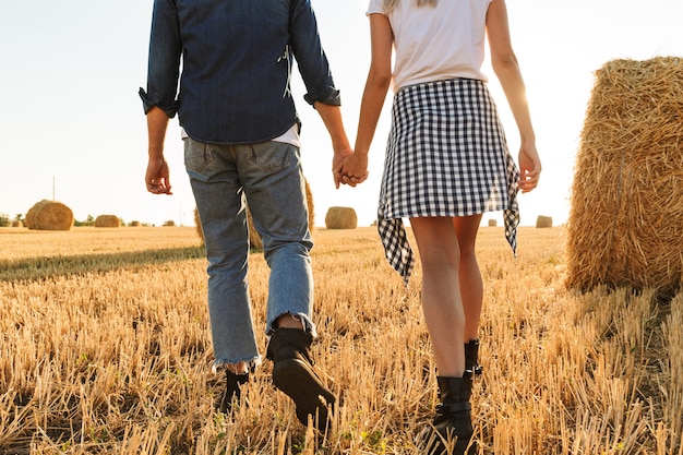 Foto ritagliata di un ragazzo e una ragazza che camminano attraverso il campo dorato con un mucchio di mucchi di fieno, durante la giornata di sole