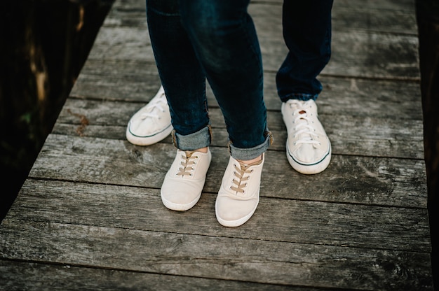 Фото Обрезанное фото молодые ноги в кроссовках, супружеская пара, муж и жена на деревянном мосту у озера. вид сзади пара, стоящая на пирсе. нижняя половина.