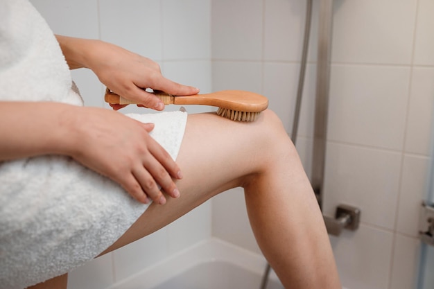 Cropped photo of a young woman in white towel doing body lymphatic drainage massage with dry wooden brush with natural bristles in bathroom at home Anticellulite exercises