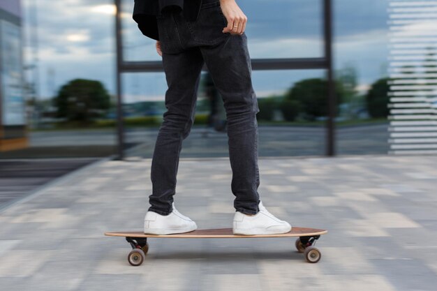 Cropped picture of male in black jeans and white sneakers riding a longboard in urban area