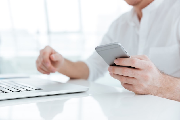 Cropped picture of young man dressed in white shirt using laptop computer and phone.