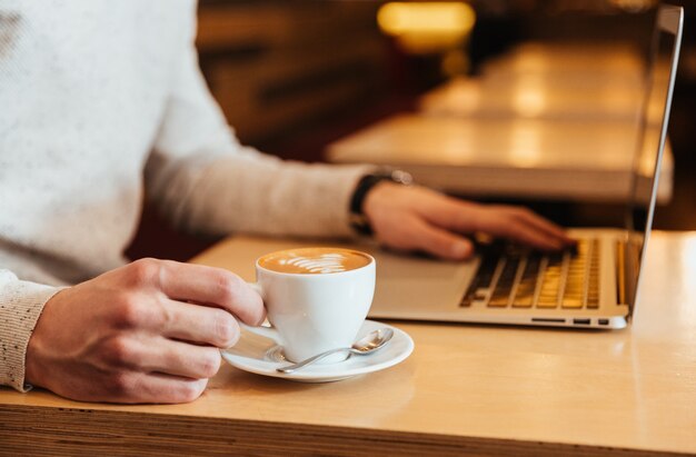 Cropped picture of young man sitting in cafe while drinking coffee and using laptop.