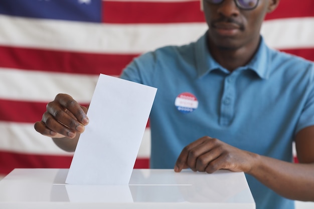 Photo cropped portrait of african-american man putting vote bulletin in ballot box while standing against american flag on election day, copy space