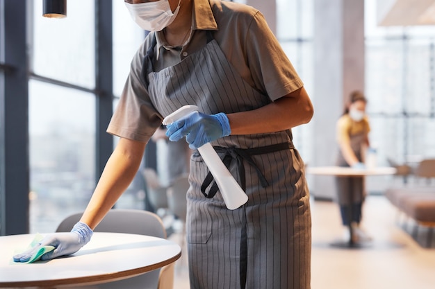 Ritratto ritagliato della lavoratrice che indossa la maschera mentre pulisce i tavoli all'interno del caffè, concetto di sicurezza covid, spazio copia