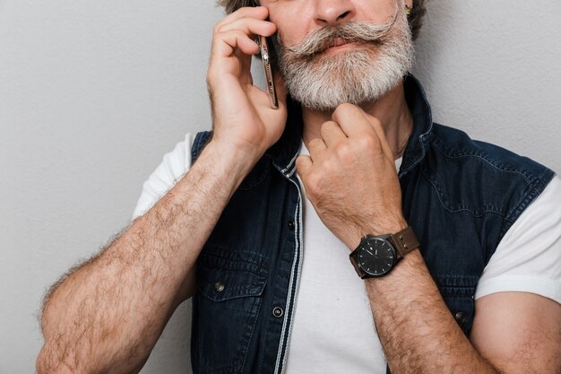 Cropped Portrait of a handsome pensive stylish mature man wearing a vest standing isolated over gray background, talking on mobile phone