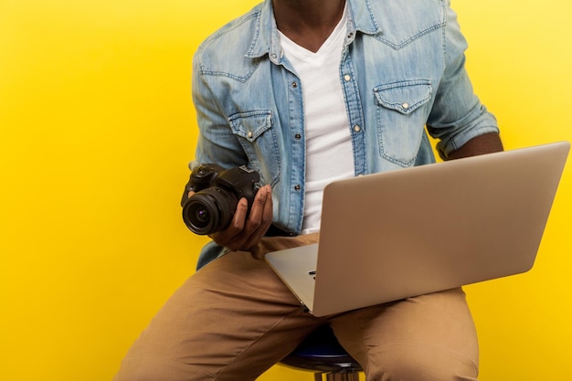 디지털 dslr 카메라와 노트북을 들고 사진 편집 앱을 사용하여 사진을 찍거나 비디오를 만들 데님 셔츠를 입은 남성 사진작가의 자른 초상화. 노란색 배경에 고립 된 스튜디오 촬영