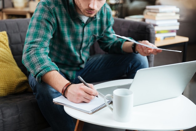 Cropped portrait of man in green shirt sitting on sofa with laptop and documents and writing by the table.