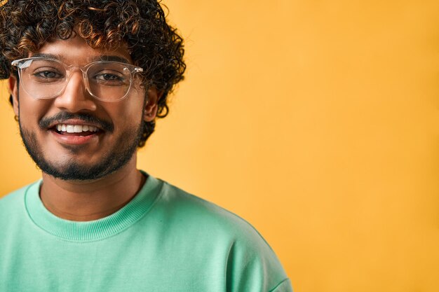 Фото Обрезанный портрет красивого кудрявого индийского мужчины в бирюзовой футболке и очках, улыбающегося и смотрящего в камеру