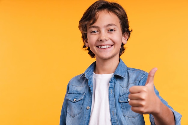 Cropped portrait of a smiling young boy showing thumb up isolated over yellow background