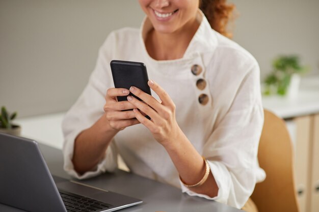 Cropped portrait of smiling young woman using smartphone at workplace while enjoying work from home, copy space