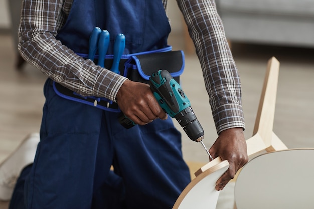 Cropped portrait of unrecognizable African-American handyman holding electric screwdriver while assembling furniture in home interior, copy space