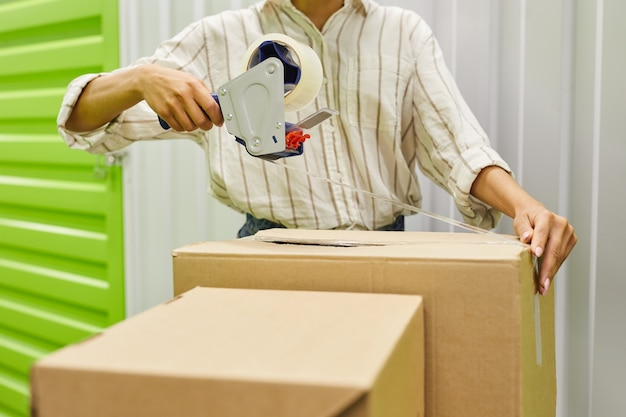 Photo cropped portrait of unrecognizable young woman packing boxes with tape gun while standing by self storage unit, copy space