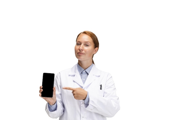 白い背景の上に分離された電話画面を指している若い女性医師のトリミングされた肖像画