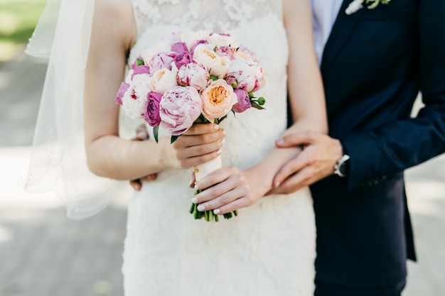 Cropped shot of beautiful bride in white wedding dress holding bouquet standing near groom who is hugging her from behind