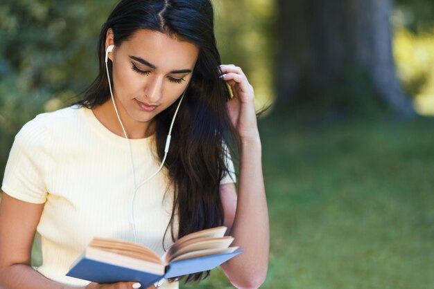 音楽を聴き、公園で本を読んでいるイヤホンで美しい白人ブルネットの若い女性のトリミングされたショット魅力的な女性のスタイリングと本のレッスンを学ぶコピースペース