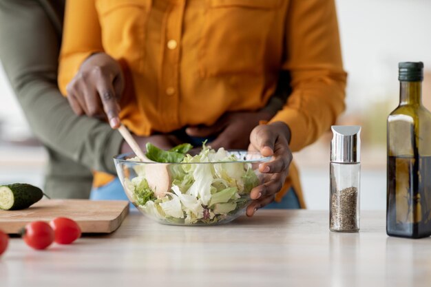 Cropped shot of black couple cooking salad together and embracing in kitchen