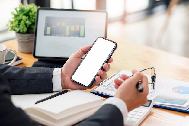 Photo cropped shot of businessman hand holding blank screeen smartphone while sitting at the table in modern office room, mock up blank screen for graphic design or product display.