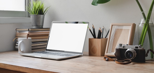 Colpo potato di area di lavoro comoda con il computer portatile dello schermo in bianco, gli articoli per ufficio e la macchina fotografica