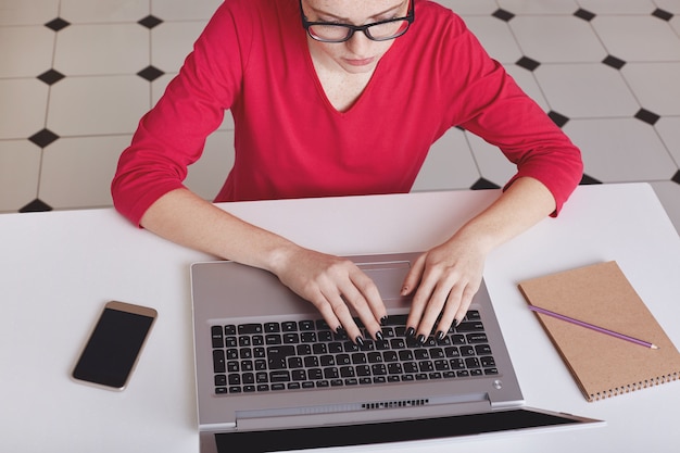 Cropped shot of concentrated female in eyewear keyboards on laptop computer.