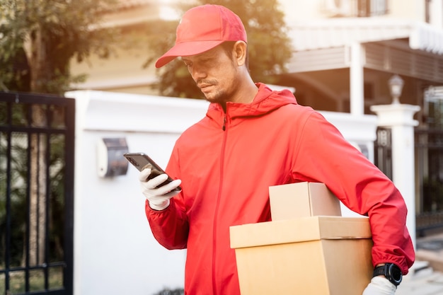 Cropped shot of a delivery man in red uniform holds postal parcel stands on street looks at cellphone checking gps address to deliver things to the customer. Delivery service during COVID-19 pandemic.