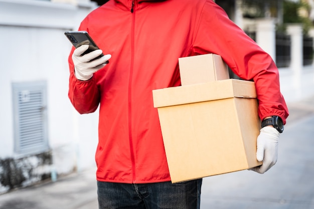 Photo cropped shot of a delivery man in red uniform holds postal parcel stands on street looks at cellphone checking gps address to deliver things to the customer. delivery service during covid-19 pandemic.