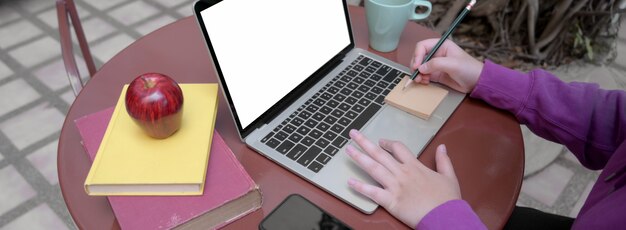 Cropped shot of female university student doing her homework with blank screen laptop and books