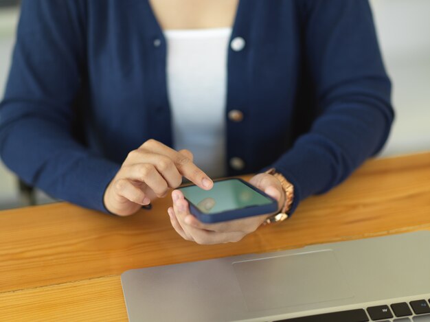 Cropped shot of female using smartphone while working with laptop on wooden desk