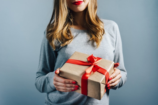 Photo cropped shot of a girl with red lips and polished nails holding gift box wrapped in craft paper and decorated with red satin ribbon