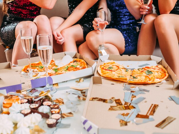 Cropped shot of ladies sitting in front of pizza in boxes, plate with sweets and glasses with sparkling wine.