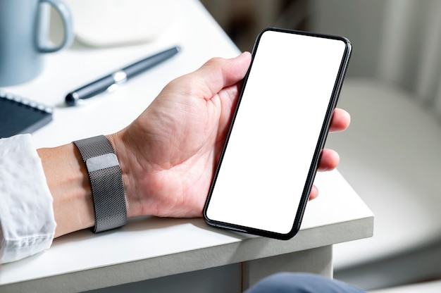 Обрезанный снимок руки человека, держащей смартфон с пустым экраном, сидя за столом.